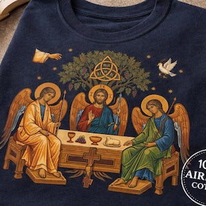 Könnte beinhalten: Marineblaues T-Shirt mit einer religiösen Illustration. Das Design zeigt drei Figuren um einen Tisch, mit einer zentralen Figur und zwei Engeln. Eine Taube, ein Baum und ein Kreuz sind ebenfalls vorhanden. Das T-Shirt hat ein rundes Etikett mit der Aufschrift "100% AIRLUME COTTON."