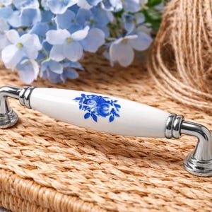 Può includere: Una maniglia per mobili in ceramica bianca con un motivo floreale blu e finiture cromate. La maniglia ovale poggia su una superficie tessuta, con fiori blu e un gomitolo di spago sullo sfondo.