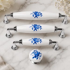 May include: Three white ceramic drawer pulls of varying lengths and one round ceramic knob, all featuring a blue floral pattern and polished chrome metal hardware. The handles have a smooth, elongated center section with metallic end caps.