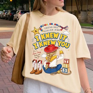 May include: A cream-colored t-shirt featuring a graphic print of a person with a blonde braid wearing a red cowboy hat, blue jeans, and white cowboy boots. The design includes text reading "I KNEW IT I KNEW YOU" and "YOU'VE GOT A FRIEN IN ME".