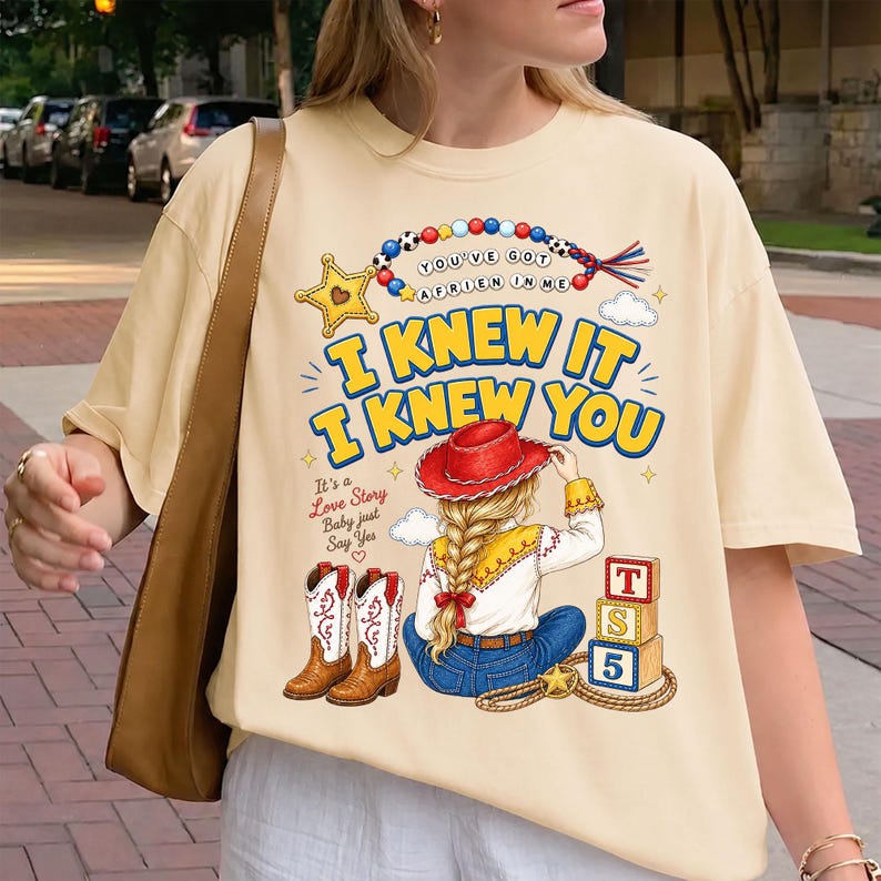 May include: A cream-colored t-shirt featuring a graphic print of a person with a blonde braid wearing a red cowboy hat, blue jeans, and white cowboy boots. The design includes text reading "I KNEW IT I KNEW YOU" and "YOU'VE GOT A FRIEN IN ME".