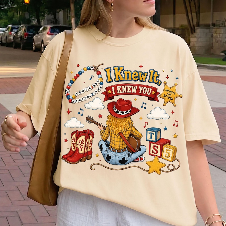May include: A cream-colored t-shirt featuring a graphic print of a cowgirl playing a guitar, red cowboy boots, a beaded bracelet with the text "YOU'VE GOT A FRIEND IN ME," and blocks labeled "T" and "S 5." The text reads "I KNEW IT, I KNEW YOU."