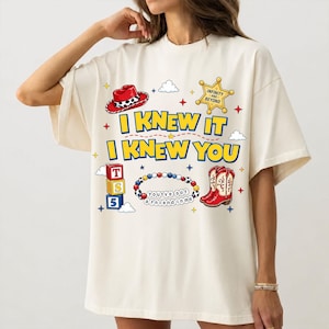 Puede incluir: Una camiseta color hueso con un estampado gr&aacute;fico que incluye el texto I KNEW IT I KNEW YOU. El dise&ntilde;o presenta un sombrero de vaquero rojo, una estrella de sheriff amarilla, bloques apilados con T S 5, una pulsera de cuentas y botas rojas.