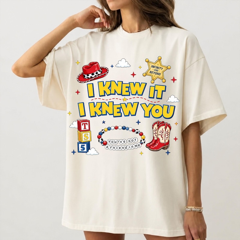 Puede incluir: Una camiseta color hueso con un estampado gr&aacute;fico que incluye el texto I KNEW IT I KNEW YOU. El dise&ntilde;o presenta un sombrero de vaquero rojo, una estrella de sheriff amarilla, bloques apilados con T S 5, una pulsera de cuentas y botas rojas.
