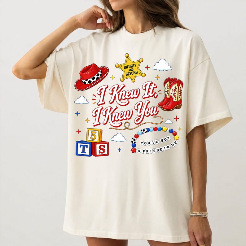 Puede incluir: Una camiseta color crema con un estampado gr&aacute;fico que incluye el texto "I Knew It, I Knew You", "Infinity and Beyond" y "You've Got a Friend in Me". El dise&ntilde;o muestra un sombrero de vaquero rojo, una estrella de sheriff amarilla, botas rojas, bloques y cuentas.