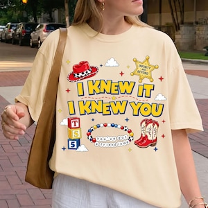 May include: A cream-colored t-shirt featuring a graphic print with a red cowboy hat, a yellow sheriff badge with the text INFINITY AND BEYOND, blue and yellow text reading I KNEW IT I KNEW YOU, alphabet blocks, a beaded bracelet, and red cowboy boots.
