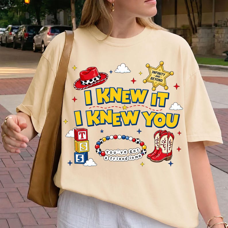 May include: A cream-colored t-shirt featuring a graphic print with a red cowboy hat, a yellow sheriff badge with the text INFINITY AND BEYOND, blue and yellow text reading I KNEW IT I KNEW YOU, alphabet blocks, a beaded bracelet, and red cowboy boots.