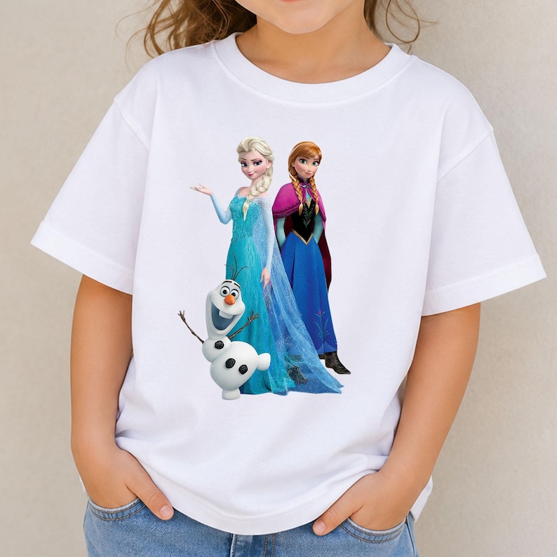 May include: A white short-sleeved t-shirt featuring a graphic print of the characters Elsa, Anna, and Olaf from the movie Frozen. Elsa wears a light blue gown, Anna wears a blue and black dress with a magenta cape, and Olaf is a white snowman.