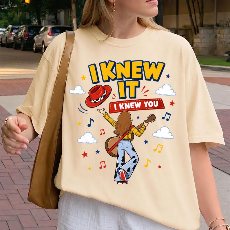 Puede incluir: Una camiseta de color crema con un gr&aacute;fico de una persona de pelo largo casta&ntilde;o tocando una guitarra ac&uacute;stica, vistiendo una camisa de cuadros amarillos y pantalones con estampado de vaca. El dise&ntilde;o incluye un sombrero de vaquero rojo, notas musicales, estrellas, nubes y el texto "I KNEW IT I KNEW YOU".
