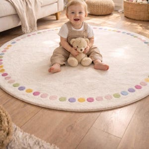 Può includere: Un tappeto rotondo bianco sporco con un bordo a pois colorati. Un bambino sorridente con una tuta beige è seduto sul tappeto, che tiene un orsacchiotto. Il tappeto è su un pavimento in legno.