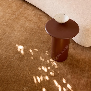 May include: A solid brown cylindrical side table with a flat circular top, topped with a small off-white ceramic bowl featuring dark speckles. The table rests on a textured brown rug with light patterns cast by sunlight across the surface.