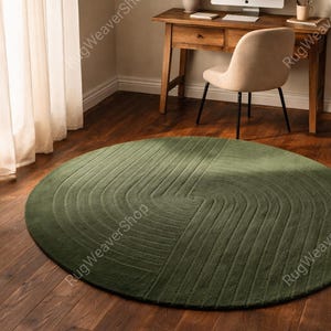 May include: A circular olive green rug featuring a textured pattern of concentric, curved lines that create a swirling effect across the surface. The rug has a dense, uniform pile and is positioned flat on a dark brown wooden floor surface.