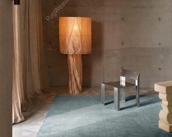 Puede incluir: Una alfombra rectangular grande de color azul sólido colocada en el suelo. La habitación cuenta con una lámpara de pie cilíndrica alta con una pantalla de tela texturizada de color claro, una silla metálica y una mesa auxiliar escultural.