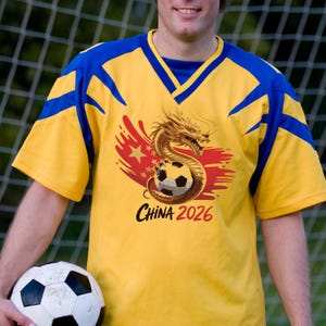 K&ouml;nnte beinhalten: Gelbes und blaues Fu&szlig;balltrikot mit einem Drachen- und Fu&szlig;ball-Design. Auf dem Trikot steht "CHINA 2026". Eine Person h&auml;lt das Trikot und einen Fu&szlig;ball.