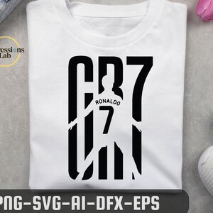 May include: A white t-shirt featuring a black graphic design of the text CR7. The design incorporates a silhouette of a soccer player wearing a jersey with the name Ronaldo and the number 7 printed on the back, integrated into the large letters.