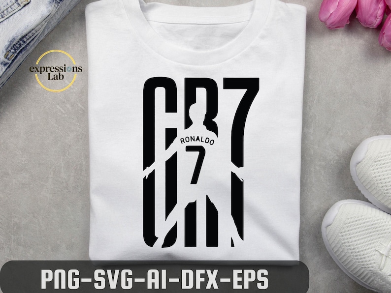 May include: A white t-shirt featuring a black graphic design of the text CR7. The design incorporates a silhouette of a soccer player wearing a jersey with the name Ronaldo and the number 7 printed on the back, integrated into the large letters.