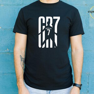 May include: A black short-sleeved t-shirt featuring a white graphic print on the chest. The design displays the text CR7 with a silhouette of a soccer player in the center, including the name Ronaldo and the number 7. The top right corner has a logo.