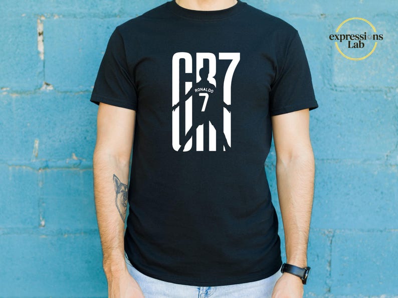 May include: A black short-sleeved t-shirt featuring a white graphic print on the chest. The design displays the text CR7 with a silhouette of a soccer player in the center, including the name Ronaldo and the number 7. The top right corner has a logo.