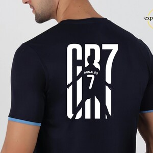 May include: A dark navy blue short-sleeved t-shirt with light blue trim on the sleeves. The back features a large white graphic print of the text CR7, incorporating a silhouette of a soccer player with the name RONALDO and the number 7 inside.