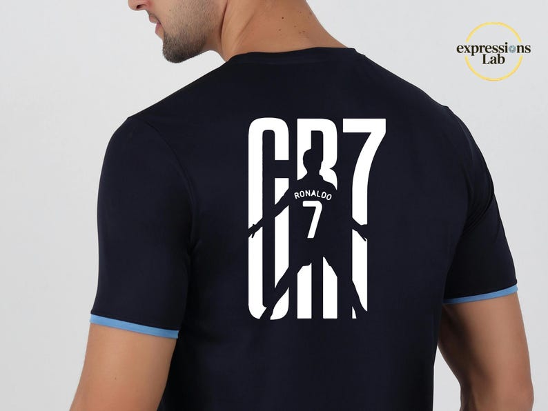 May include: A dark navy blue short-sleeved t-shirt with light blue trim on the sleeves. The back features a large white graphic print of the text CR7, incorporating a silhouette of a soccer player with the name RONALDO and the number 7 inside.