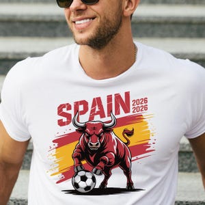K&ouml;nnte beinhalten: Wei&szlig;es T-Shirt mit einer Grafik eines roten Bullen mit einem Fu&szlig;ball, dem Wort "SPAIN" in Rot und den Zahlen "2026" zweimal. Der Bulle steht vor einem roten und gelben Hintergrund.