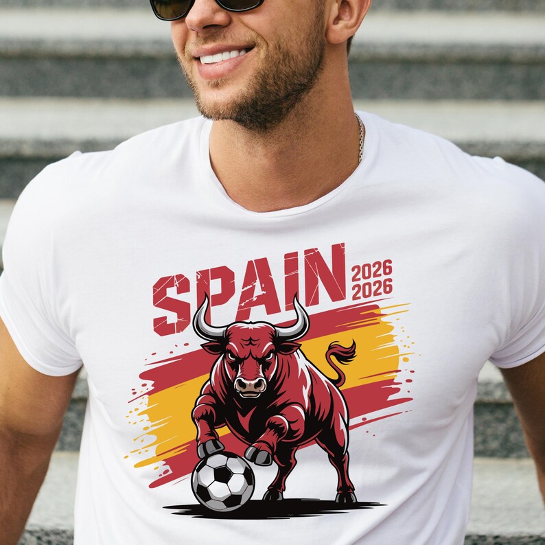 K&ouml;nnte beinhalten: Wei&szlig;es T-Shirt mit einer Grafik eines roten Bullen mit einem Fu&szlig;ball, dem Wort "SPAIN" in Rot und den Zahlen "2026" zweimal. Der Bulle steht vor einem roten und gelben Hintergrund.