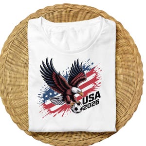 K&ouml;nnte beinhalten: Wei&szlig;es T-Shirt mit einer Grafik eines Adlers, der einen Fu&szlig;ball h&auml;lt, vor einem Hintergrund aus Rot, Wei&szlig; und Blau. Der Text "USA 2026" ist in Schwarz dargestellt und repr&auml;sentiert ein Sportereignis.