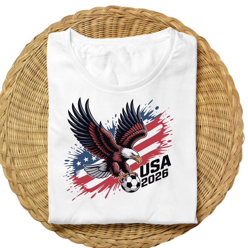 K&ouml;nnte beinhalten: Wei&szlig;es T-Shirt mit einer Grafik eines Adlers, der einen Fu&szlig;ball h&auml;lt, vor einem Hintergrund aus Rot, Wei&szlig; und Blau. Der Text "USA 2026" ist in Schwarz dargestellt und repr&auml;sentiert ein Sportereignis.
