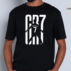 May include: A black short-sleeved t-shirt featuring a white graphic print on the chest. The design displays the text CR7 with a silhouette of a soccer player in the center, including the name RONALDO and the number 7. The logo expressions Lab is visible.