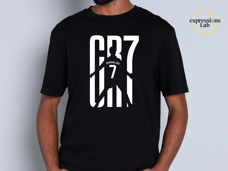 May include: A black short-sleeved t-shirt featuring a white graphic print on the chest. The design displays the text CR7 with a silhouette of a soccer player in the center, including the name RONALDO and the number 7. The logo expressions Lab is visible.