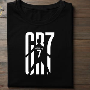 May include: A black short-sleeved t-shirt featuring a white graphic print. The design displays the text CR7 in large block letters, with a silhouette of a soccer player wearing a jersey with the name RONALDO and the number 7 integrated into the center.
