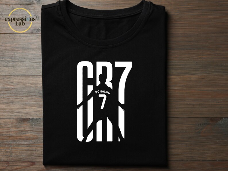 May include: A black short-sleeved t-shirt featuring a white graphic print. The design displays the text CR7 in large block letters, with a silhouette of a soccer player wearing a jersey with the name RONALDO and the number 7 integrated into the center.