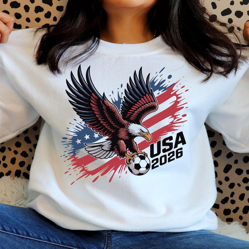 K&ouml;nnte beinhalten: Wei&szlig;es Sweatshirt mit einer Grafik eines Wei&szlig;kopfseeadlers, der einen Fu&szlig;ball h&auml;lt. Das Design enth&auml;lt rote, wei&szlig;e und blaue Elemente mit dem Text "USA 2026". Die Fl&uuml;gel des Adlers sind ausgebreitet.