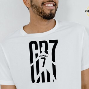 May include: A white short-sleeved t-shirt featuring a black graphic print on the chest. The design displays the text CR7 with a silhouette of a soccer player in the center, including the name RONALDO and the number 7 printed on the player's back.