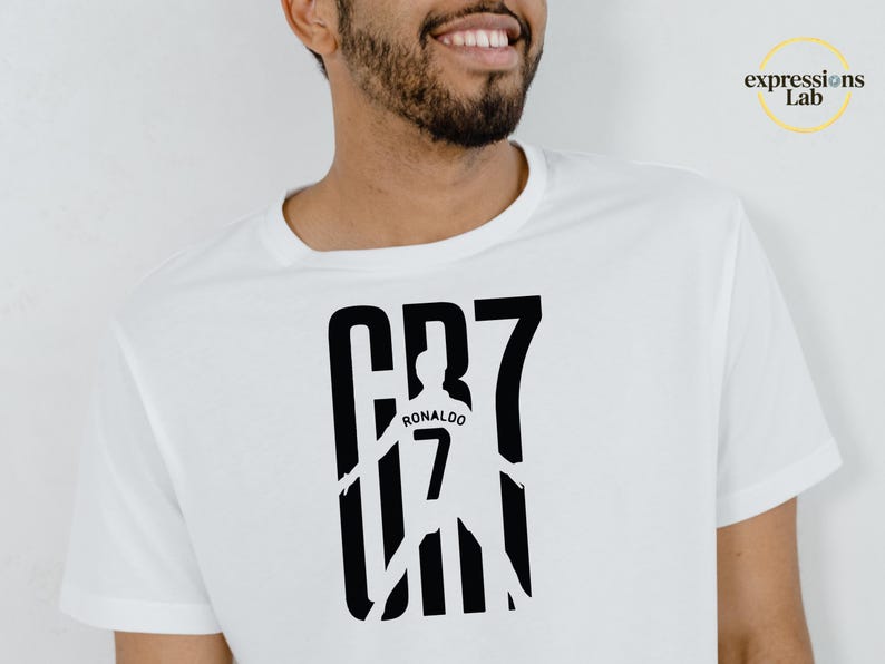 May include: A white short-sleeved t-shirt featuring a black graphic print on the chest. The design displays the text CR7 with a silhouette of a soccer player in the center, including the name RONALDO and the number 7 printed on the player's back.