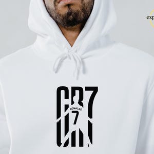 May include: A white hooded sweatshirt featuring a black graphic print on the chest. The design displays the text CR7 with a silhouette of a soccer player wearing a jersey with the number 7 and the name Ronaldo. The logo Expressions Lab is visible.