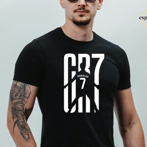 May include: A black short-sleeved t-shirt featuring a large white graphic on the chest. The design displays the text CR7 in a bold font, with a silhouette of a soccer player wearing a jersey with the name Ronaldo and the number 7 centered within.
