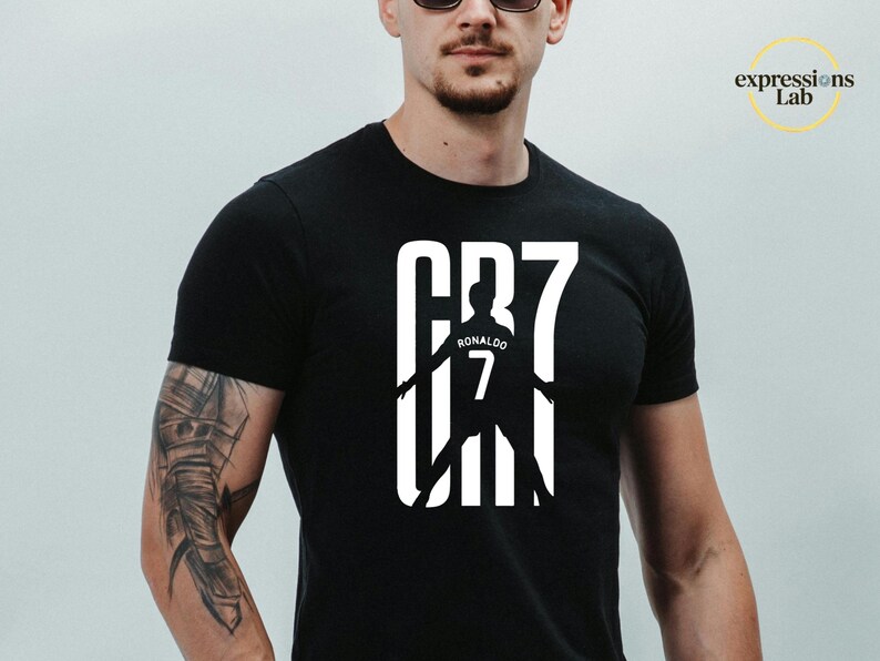 May include: A black short-sleeved t-shirt featuring a large white graphic on the chest. The design displays the text CR7 in a bold font, with a silhouette of a soccer player wearing a jersey with the name Ronaldo and the number 7 centered within.