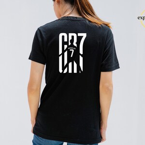 May include: A black short-sleeved t-shirt featuring a white graphic on the back. The design displays the text CR7 with a silhouette of a soccer player wearing a jersey with the name Ronaldo and the number 7 printed in the center of the graphic.