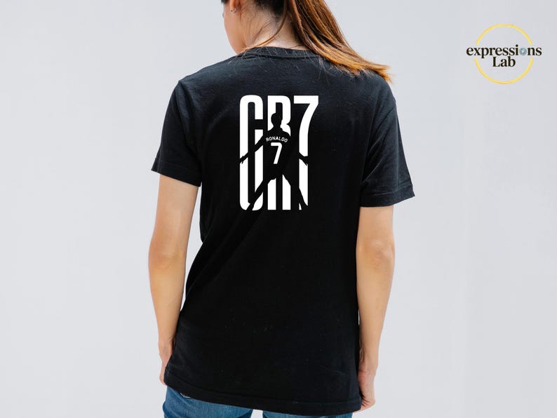 May include: A black short-sleeved t-shirt featuring a white graphic on the back. The design displays the text CR7 with a silhouette of a soccer player wearing a jersey with the name Ronaldo and the number 7 printed in the center of the graphic.