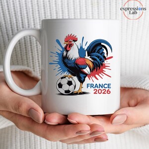 K&ouml;nnte beinhalten: Wei&szlig;er Keramikbecher mit einer farbenfrohen Illustration eines Hahns mit Fu&szlig;ball, blauen und roten Farbspritzern und dem Text "FRANCE 2026". Der Becher hat einen gro&szlig;en, gebogenen Henkel.