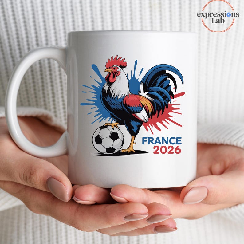 K&ouml;nnte beinhalten: Wei&szlig;er Keramikbecher mit einer farbenfrohen Illustration eines Hahns mit Fu&szlig;ball, blauen und roten Farbspritzern und dem Text "FRANCE 2026". Der Becher hat einen gro&szlig;en, gebogenen Henkel.
