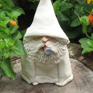 May include: A ceramic garden gnome statue with a long white beard, a pointed hat, and a cigar in its mouth. The gnome is wearing a white robe and is sitting on a tree stump. The background features green foliage and orange flowers.