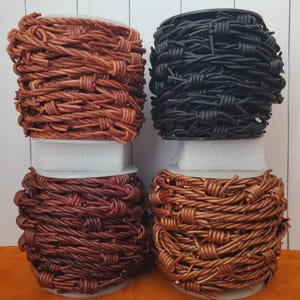 May include: Four spools of decorative barbed wire in various colors: light brown, black, dark brown, and copper. Each spool is tightly wound and sits on a white base, with an orange background and a white wooden backdrop.