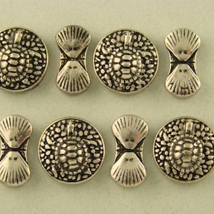 May include: A collection of eight silver-toned metal buttons arranged in two rows. Four buttons are circular with a raised turtle motif on a textured background, and four buttons are shaped like an hourglass with a ribbed shell-like pattern.
