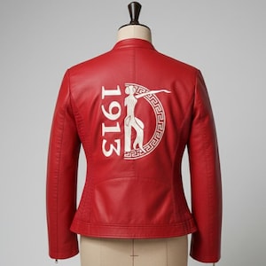 May include: Red faux leather jacket with a stand-up collar. The back features a white embroidered design with the year "1913" and a stylized figure within a decorative border. The jacket has a fitted silhouette and long sleeves.