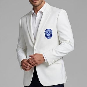 Könnte beinhalten: Ein weißer Blazer mit einem blauen Emblem auf der Brust. Der Blazer hat ein fallendes Revers, einen Zwei-Knopf-Verschluss und eine Brusttasche. Das Emblem ist ein Wappen mit Text und einem Rand. Der Blazer wird über einem weißen Hemd getragen.