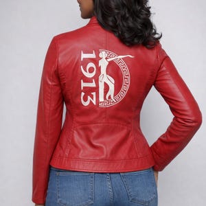 May include: A red leather jacket with a stand-up collar and a tailored fit. The back of the jacket features white embroidered text and a graphic design. The jacket is worn with blue jeans.