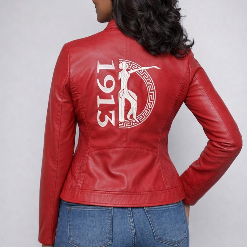 May include: A red leather jacket with a stand-up collar and a tailored fit. The back of the jacket features white embroidered text and a graphic design. The jacket is worn with blue jeans.