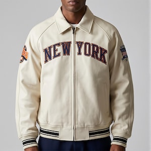 May include: Cream-colored jacket with the text "NEW YORK" in navy blue and orange lettering across the chest. The jacket has a collared neckline, a zipper closure, and patches on the sleeves. The cuffs and hem feature black and white striped detailing.